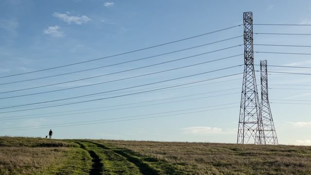 Aukštos įtampos elektros linija virš žolėto lauko, tolumoje einantis žmogus